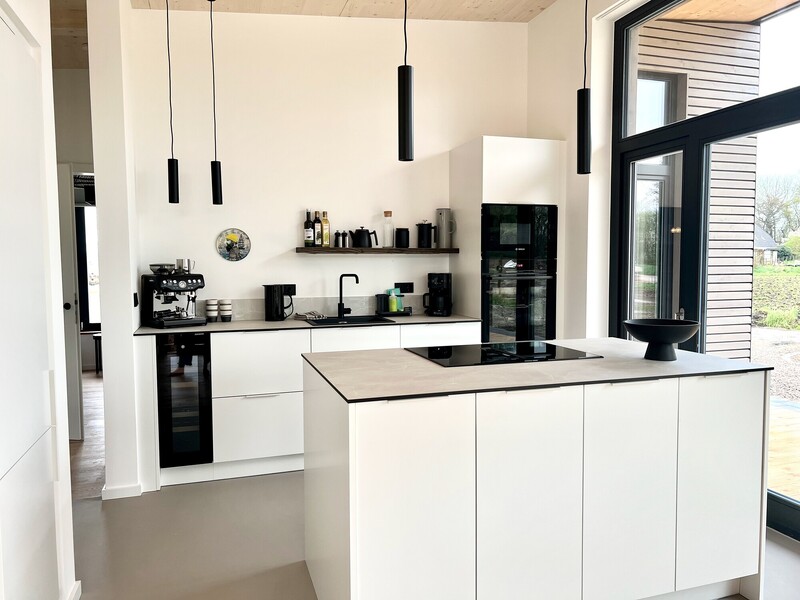 Küche im StrandHaus mit weißen Fronten, Kochinsel, schwarzen Pendelleuchten und Einbaugeräten