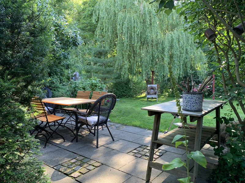 Idyllische Gartenterrasse mit Holztisch, Stühlen und Beistelltisch unter Trauerweide, umgeben von viel Grün