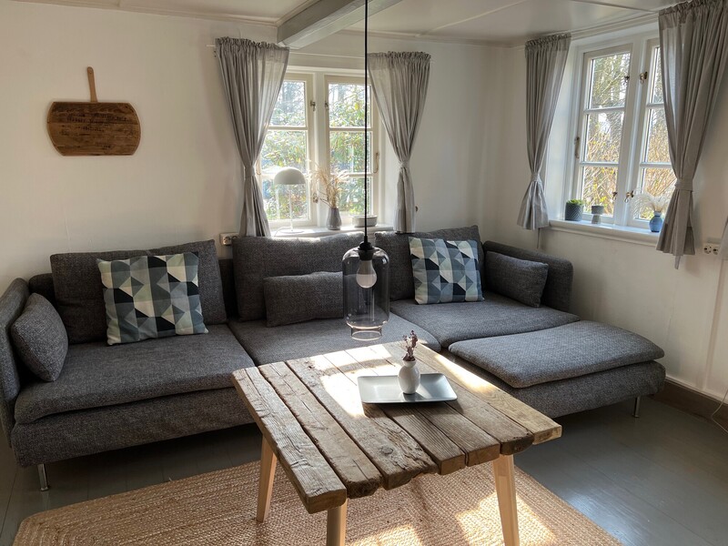 Gemütliches Wohnzimmer mit grauem Ecksofa, rustikalem Holz-Couchtisch und Sprossenfenstern mit Blick ins Grüne