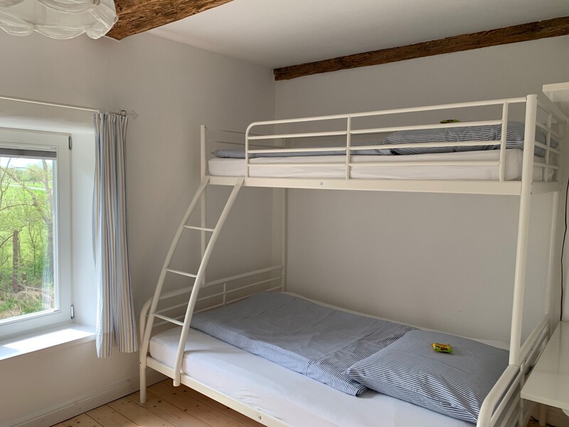 Kinderzimmer mit weißem Etagenbett, blau-weiß gestreifter Bettwäsche, rustikalem Holzdeckenbalken und Fenster mit Blick ins Grüne