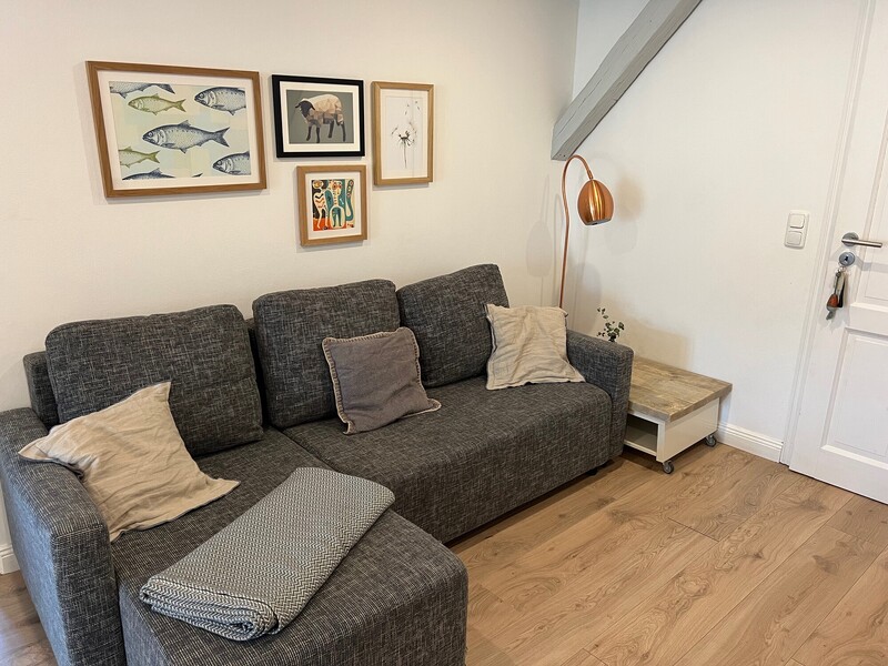 Wohnbereich mit grauem Ecksofa, Kissen, Kupfer-Stehlampe und gerahmten Bildern an der Wand; sichtbarer Deckenbalken und Dielenboden
