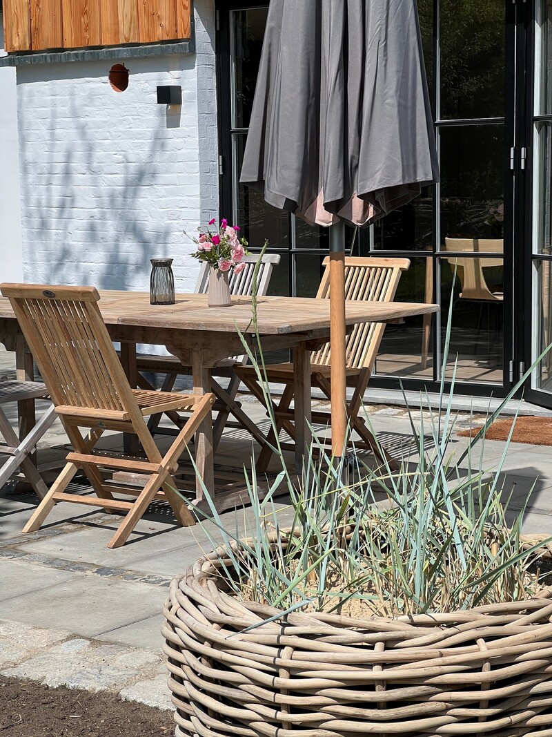 Sonnige Terrasse mit Holztisch und -stühlen, Sonnenschirm und bepflanztem Korb vor der weißen Hauswand mit Sprossenfenstern