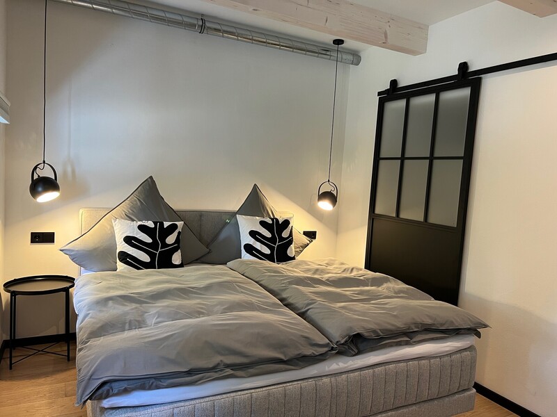 Modernes Schlafzimmer mit Doppelbett, grauer Bettwäsche und schwarz-weißen Kissen, Pendelleuchten und schwarzer Schiebetür im Loft-Stil