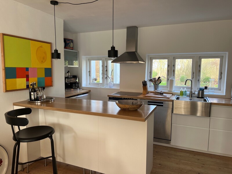 Offene Küche mit Kücheninsel und Holzarbeitsplatte, Barhockern, Edelstahlspüle und -dunstabzug sowie farbenfrohem Bild an der Wand; Fenster mit Blick ins Grüne