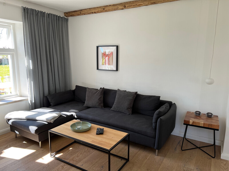 Wohnzimmer im Haus NordseeStil mit dunklem Ecksofa, Holzcouchtisch, Beistelltisch und Holzbalken an der Decke