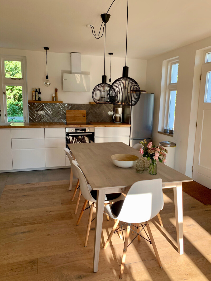 Küche mit Essplatz im Haus NordseeStil, Holztisch, weißen Stühlen, weißer Einbauküche und schwarzen Pendelleuchten