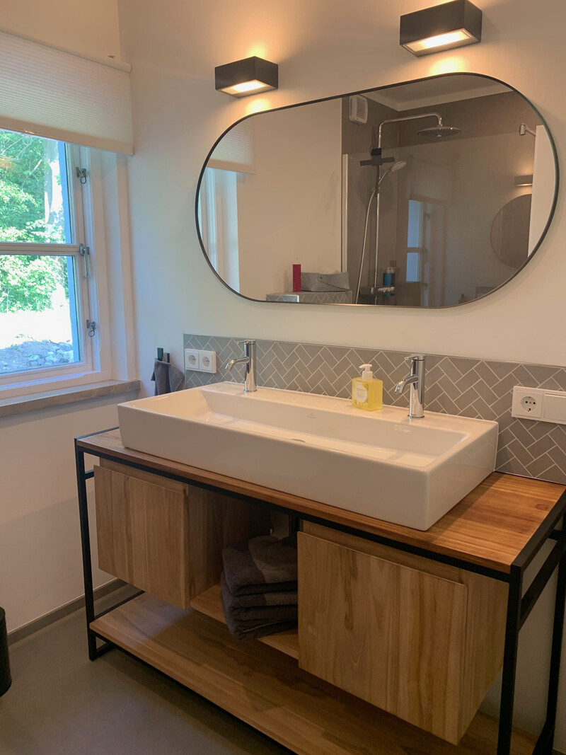 Badezimmer im Haus NordseeStil mit Doppelwaschbecken, Holz-Waschtisch, großem Spiegel und Dusche im Hintergrund