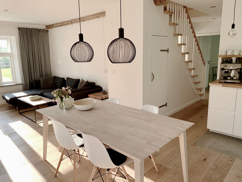 Offener Wohn-Essbereich im Haus NordseeStil mit Holzesstisch, weißen Stühlen, schwarzen Pendelleuchten und offener Küche