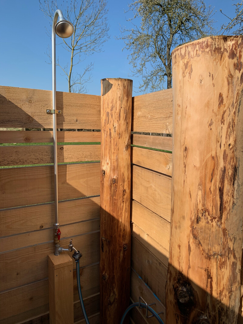 Außendusche im Haus NordseeStil mit Duschkopf, Holzverkleidung und Baumstämmen unter blauem Himmel