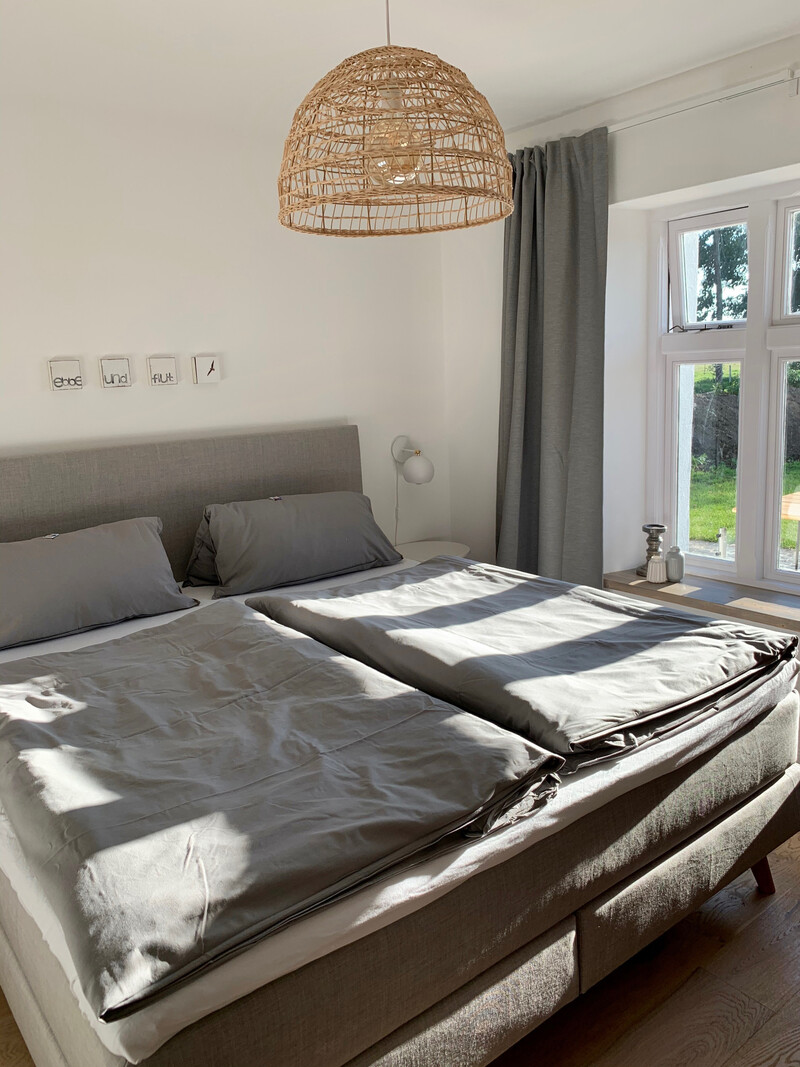 Schlafzimmer im Haus NordseeStil mit Boxspringbett, grauer Bettwäsche, Rattan-Pendelleuchte und Fenster mit Gartenblick