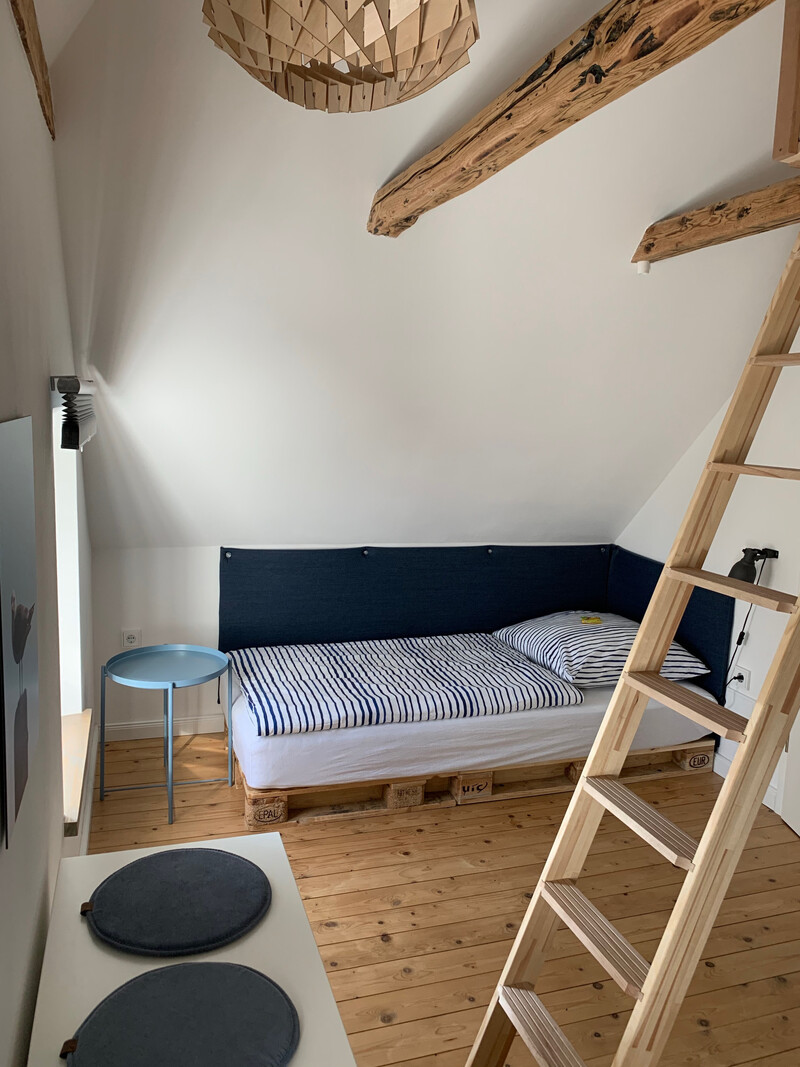 Dachzimmer im Haus NordseeStil mit Palettenbett, blau-weißer Bettwäsche, Holzleiter, Beistelltisch und Holzbalken