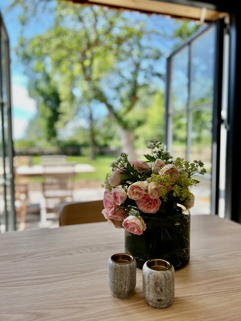 Blick vom Esstisch im Haus Scheune mit Blumenstrauß durch offene Fenstertüren auf Terrasse und Garten