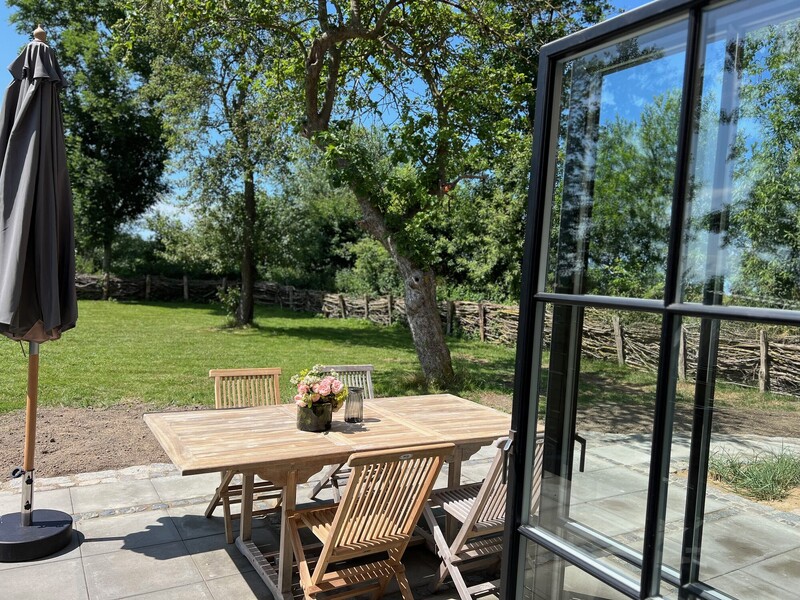 Terrasse vom Haus Scheune mit Holztisch, Stühlen, Sonnenschirm, Fenstertür und Blick auf Garten und Bäume