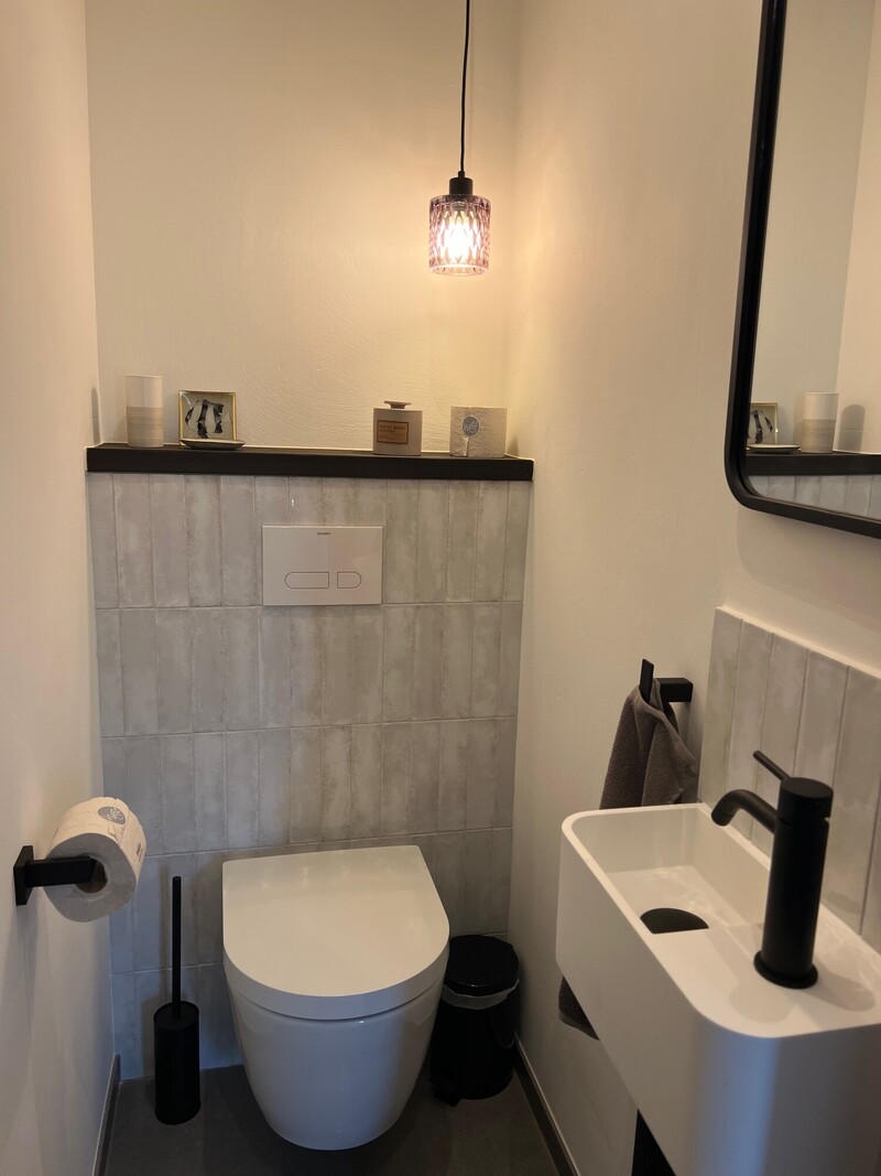Gäste-WC im Haus Scheune mit Wand-WC, kleinem Waschbecken, Spiegel, Pendelleuchte und grauen Fliesen