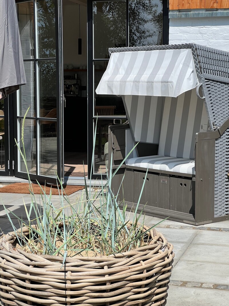 Terrasse vom Haus Scheune mit grau-weißem Strandkorb, Pflanzkorb mit Gräsern und schwarzen Fenstertüren