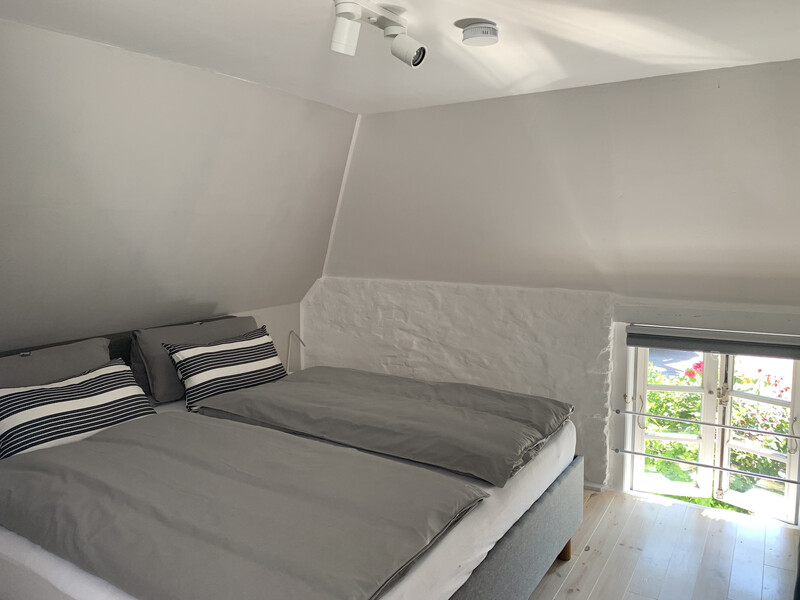 Schlafzimmer der Reetdachkate 1813 mit Doppelbett, grauer Bettwäsche, Dachschräge und Fenster mit Gartenblick