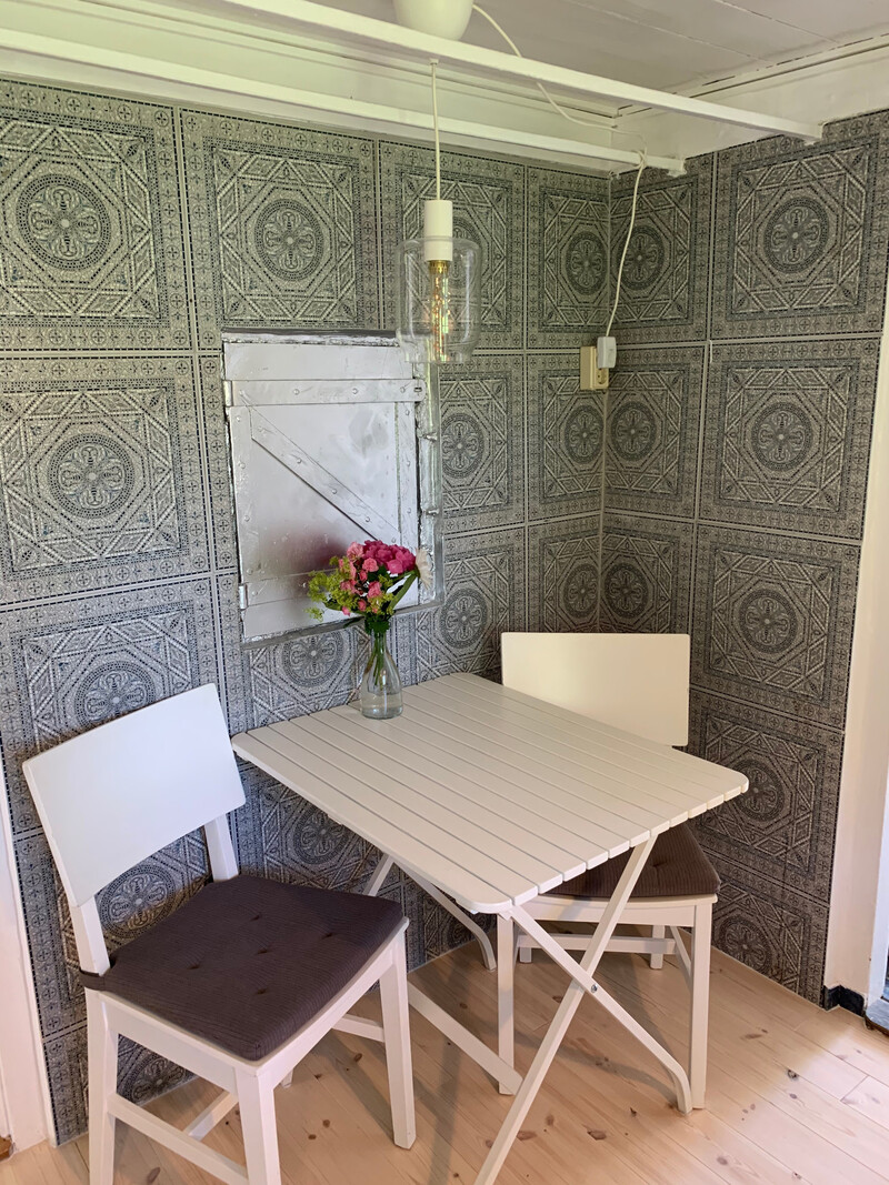 Essecke der Reetdachkate 1813 mit weißem Tisch, zwei Stühlen, gemustertem Wandbelag und Blumen als Deko