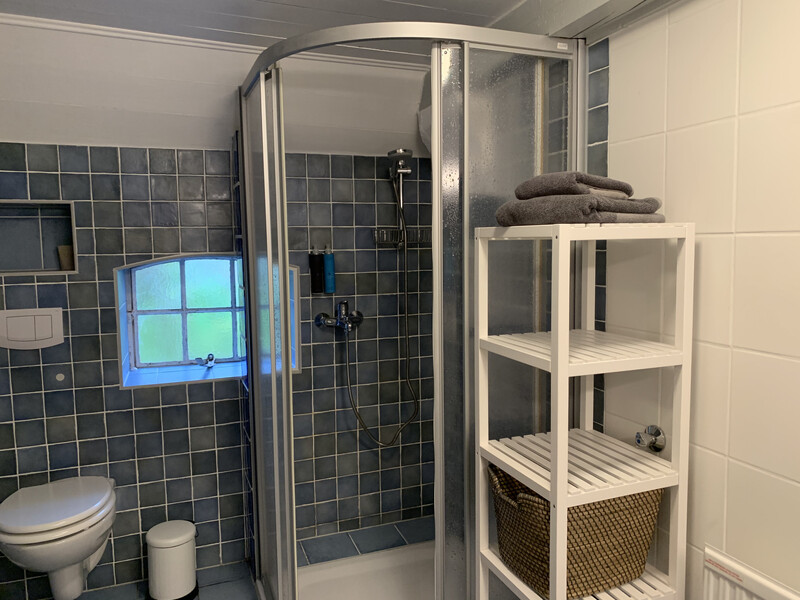Badezimmer der Reetdachkate 1813 mit Eckdusche, WC, blauen Fliesen, Regalfach mit Handtüchern und Fenster