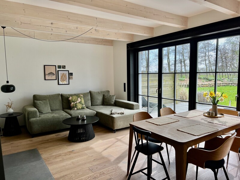 Heller Wohn-Essbereich eines Ferienhauses im Warfthof Wollatz mit grünem Ecksofa, Holzbalkendecke, Esstisch aus Holz und bodentiefen schwarzen Fenstertüren mit Blick auf Terrasse und Garten